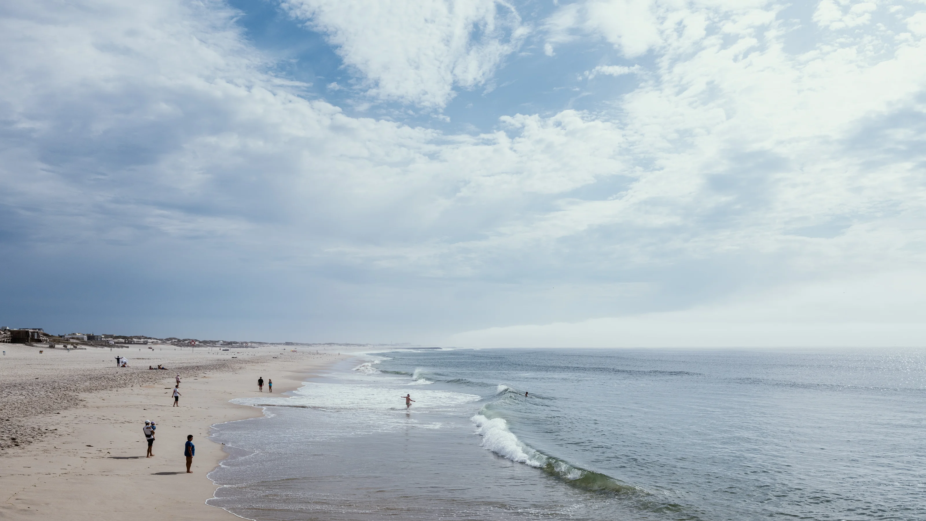 A sandy beach with people walking along the shore under a cloudy sky.