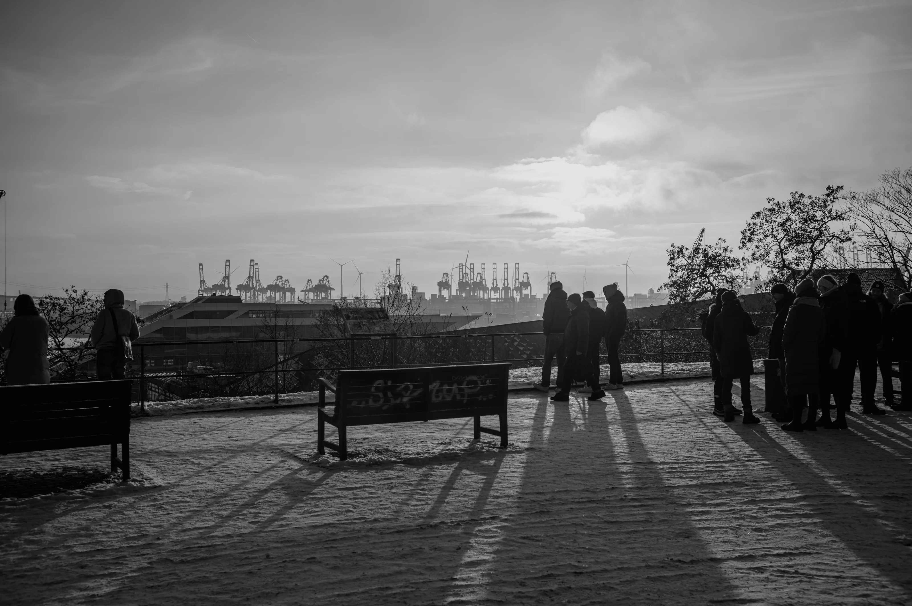 Silhouetted figures stand on a snowy path overlooking a port with cranes at sunset.