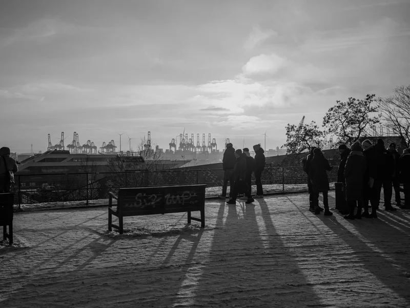 Silhouetted figures stand on a snowy path overlooking a port with cranes at sunset.
