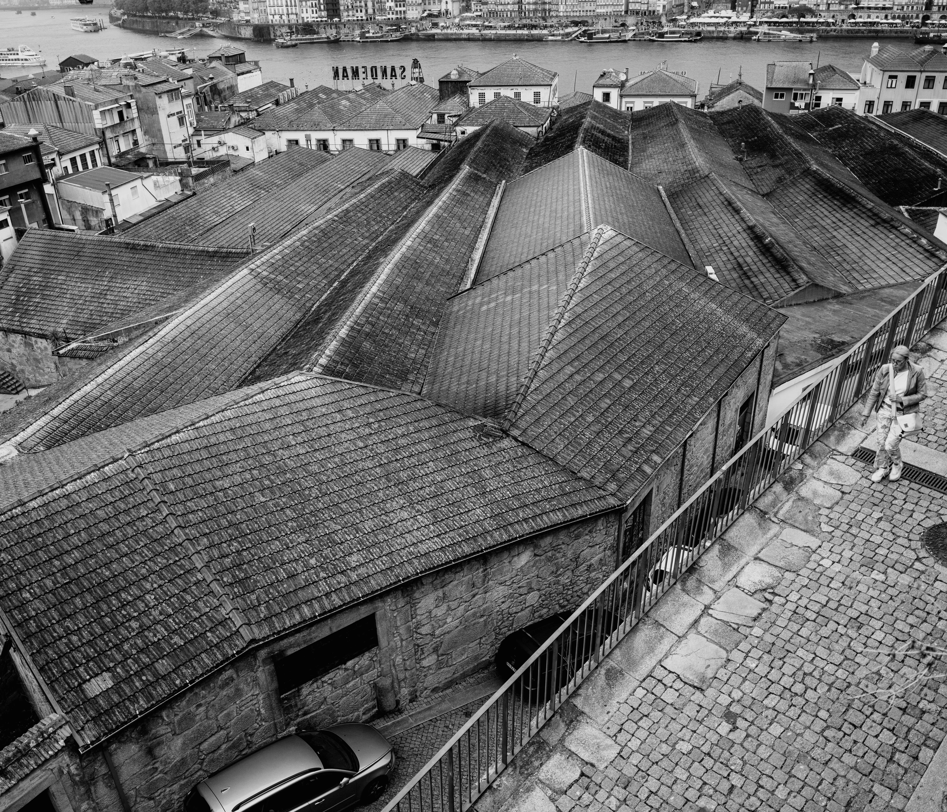 Black and white photo of tiled rooftops descending towards a river, with a person walking on a cobblestone path.