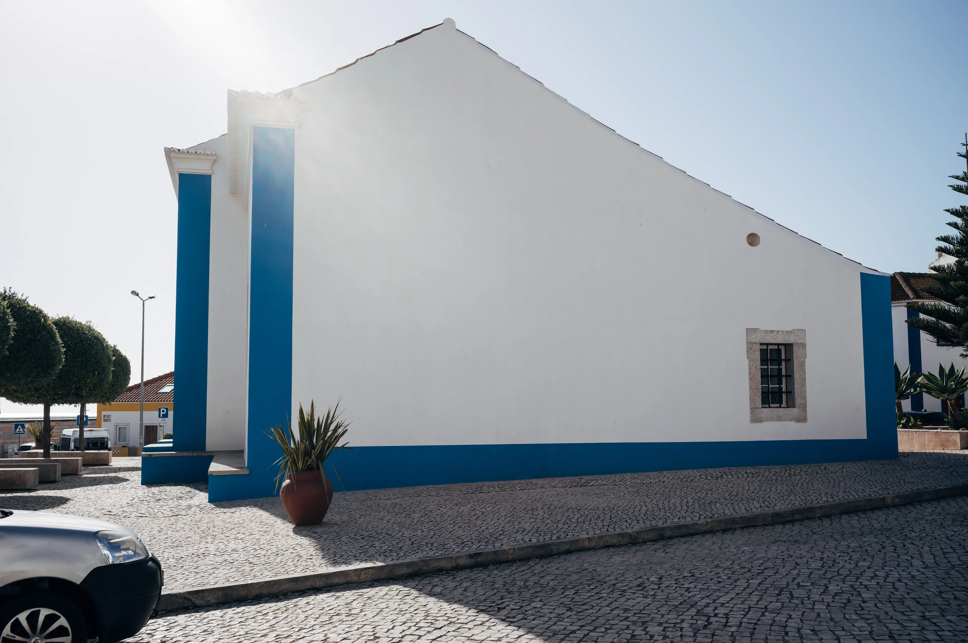 White building with blue accents under a clear sky, with sunlight creating shadows on the cobblestone ground.