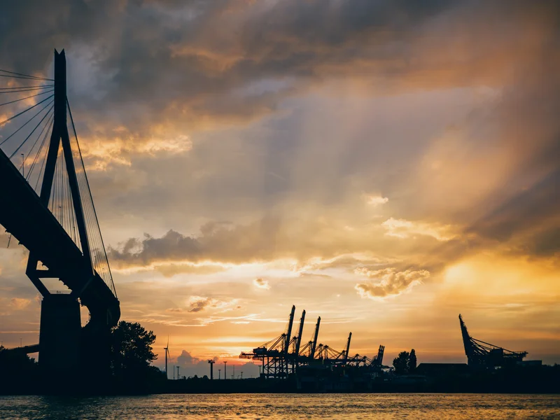 Sunset view of a bridge and port cranes silhouetted against a cloudy sky.