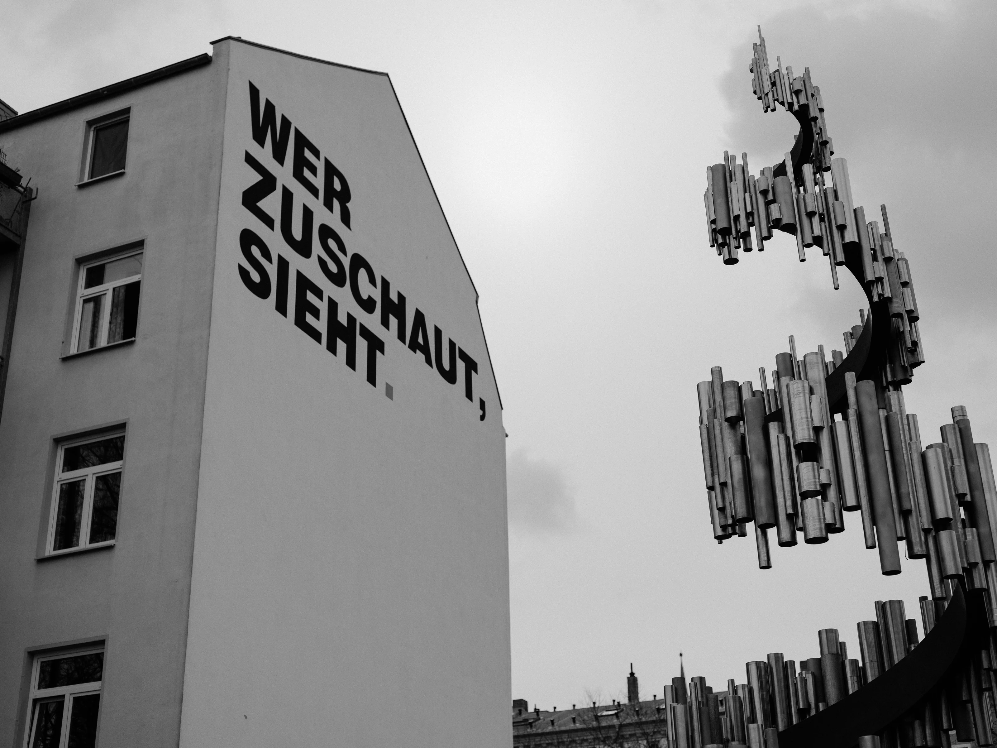 Black and white photo of a building with bold text 'WER ZUSCHAUT, SIEHT.' and a metallic sculpture.