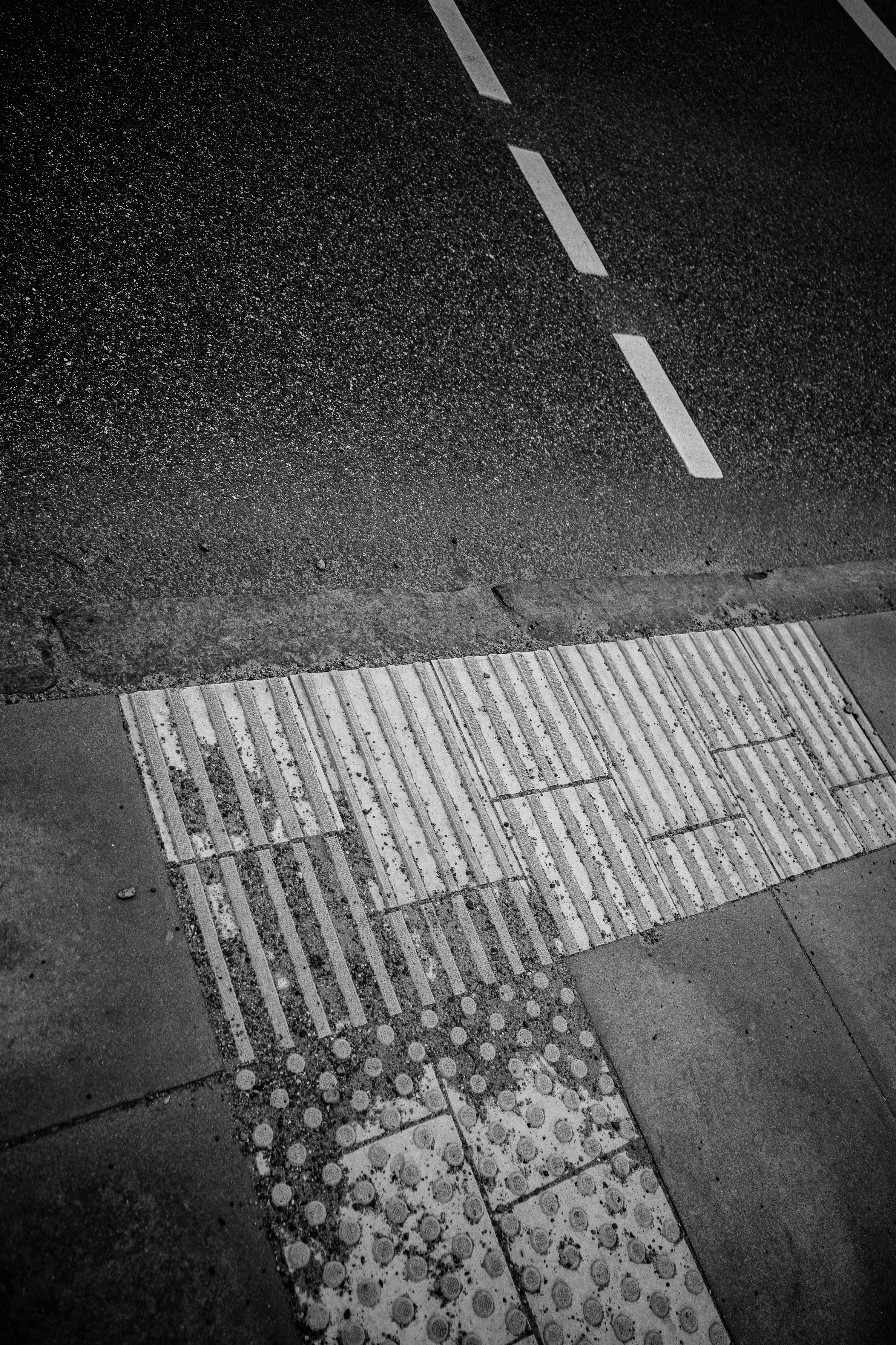 Black and white photo of a textured sidewalk edge with tactile paving next to a road with dashed lines.
