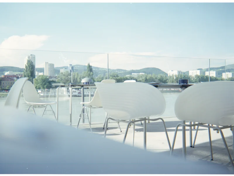 Outdoor seating area with white chairs and a glass railing overlooking a river and cityscape.