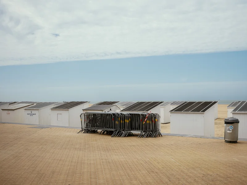 A sandy beach with several white cabins and metal barriers, under a partly cloudy sky.