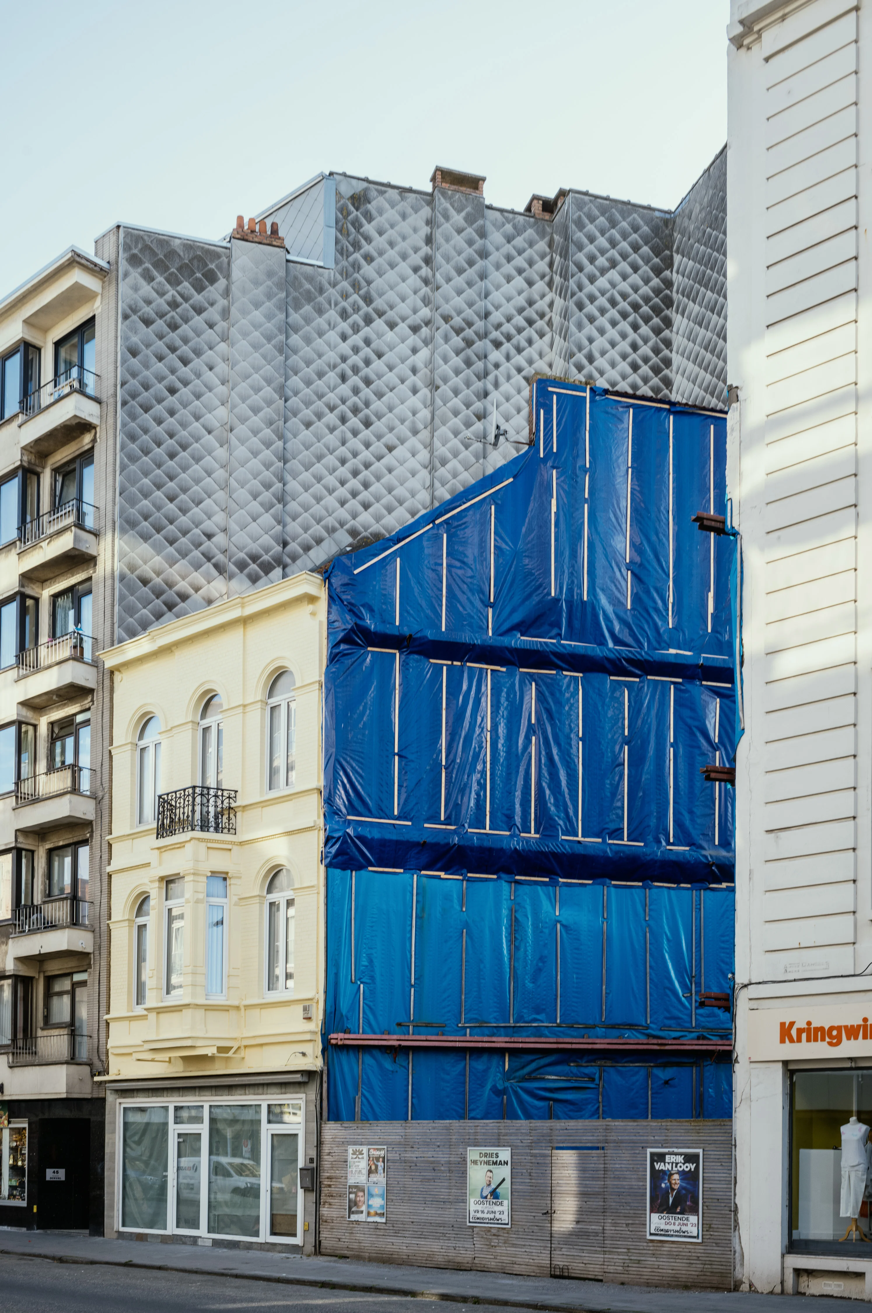 A building facade under construction with blue tarpaulin covering next to a yellow and a white building.