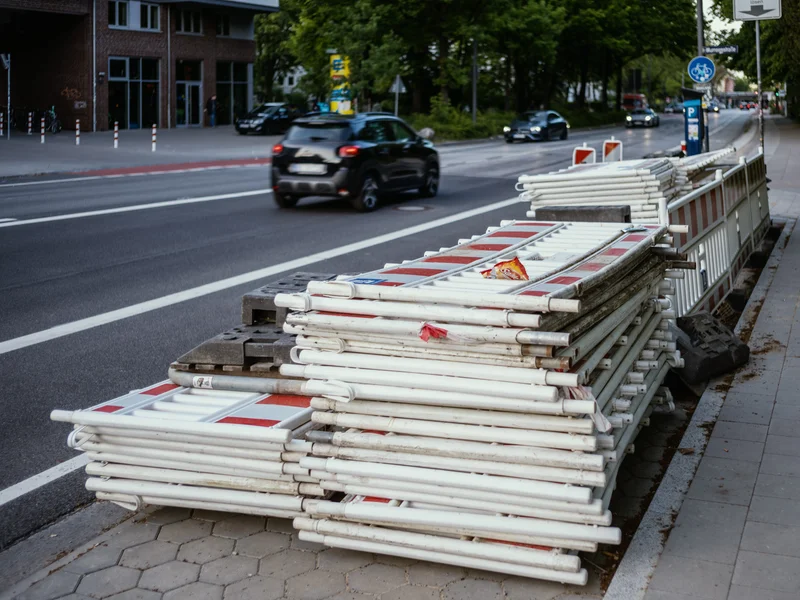 Stack of white and red construction barriers on a sidewalk beside a road with passing cars.