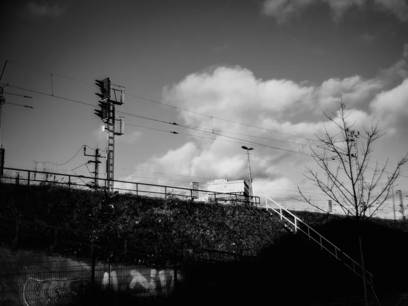 Black and white photo of a railway signal and power lines with stairs leading up a grassy slope against a cloudy sky.
