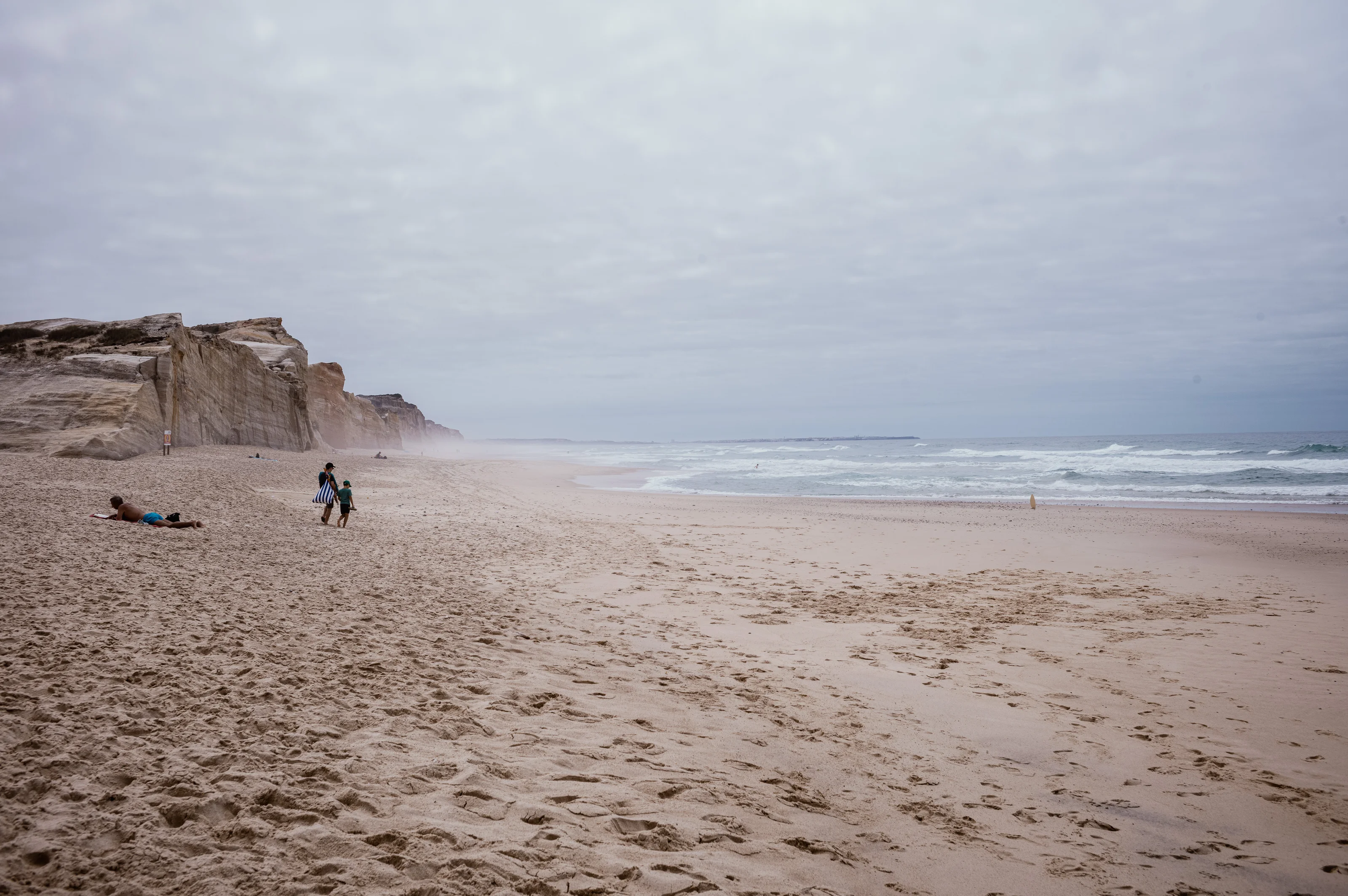 People walking along a wide, sandy beach with cliffs in the distance.