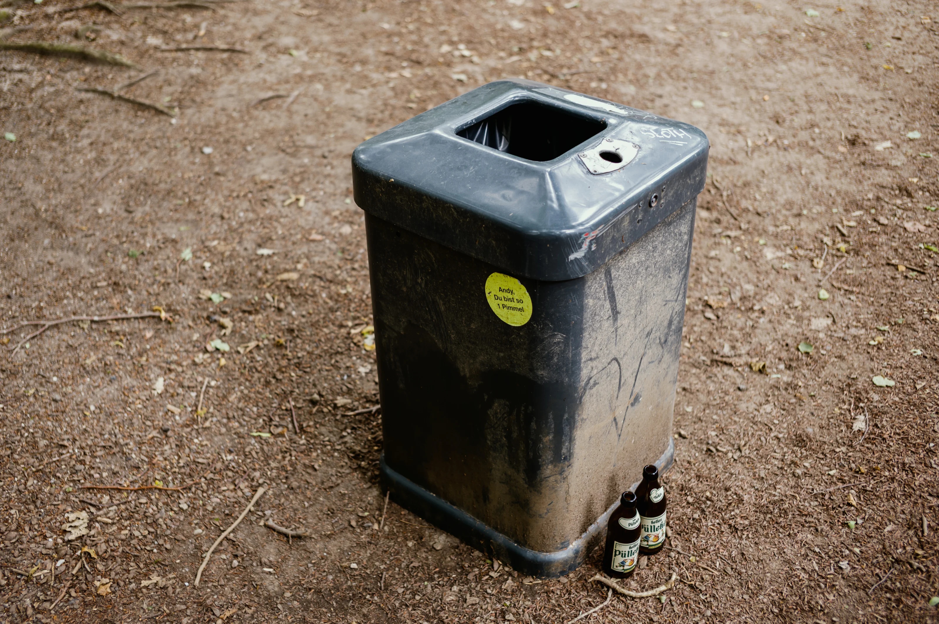 Gray trash bin with two glass bottles beside it on a dirt ground.