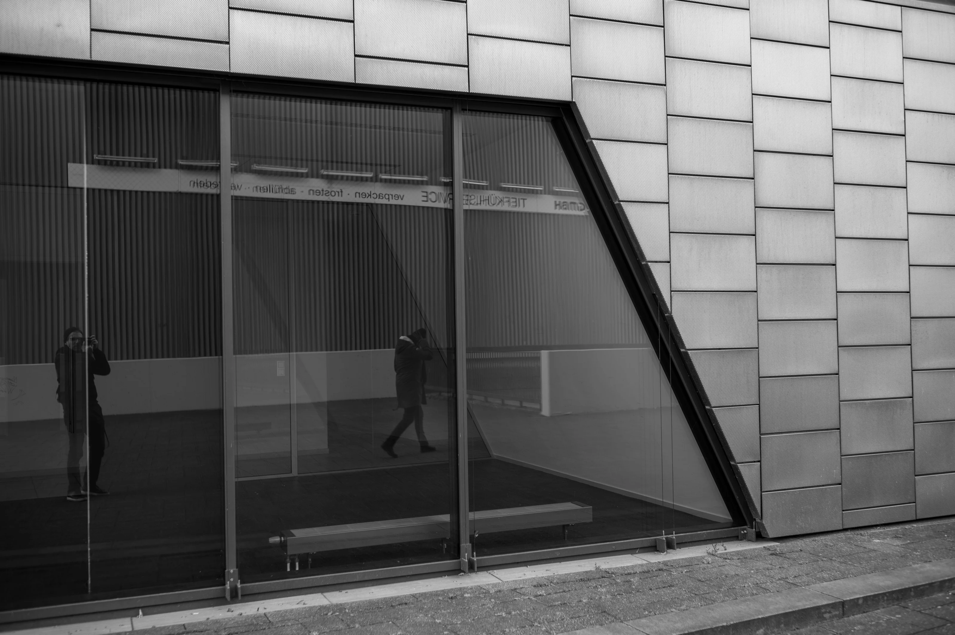 Black and white photo of a building facade with reflective windows and a person walking, reflected in the glass.