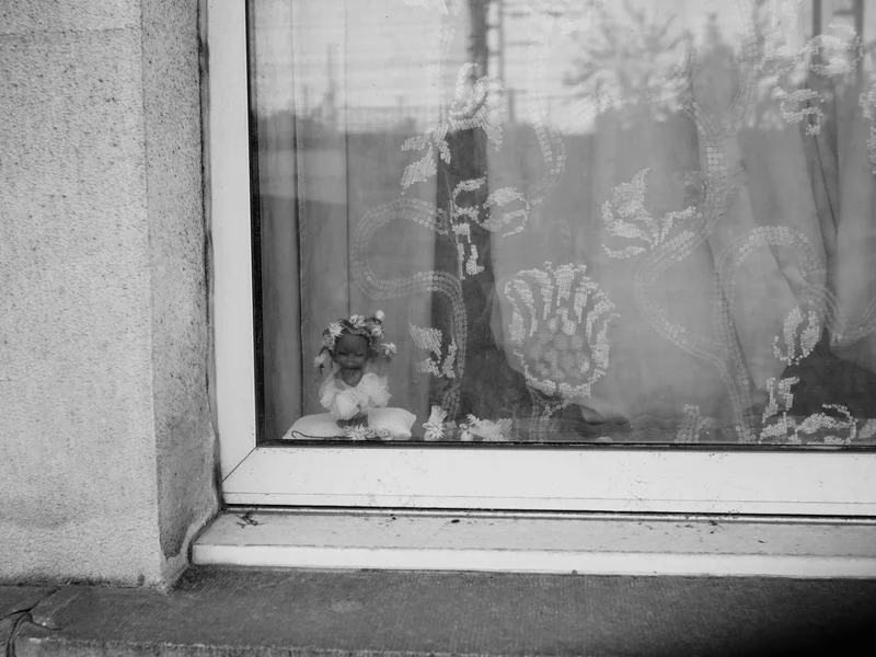 A small doll with a floral crown is visible through a lace curtain in a window.