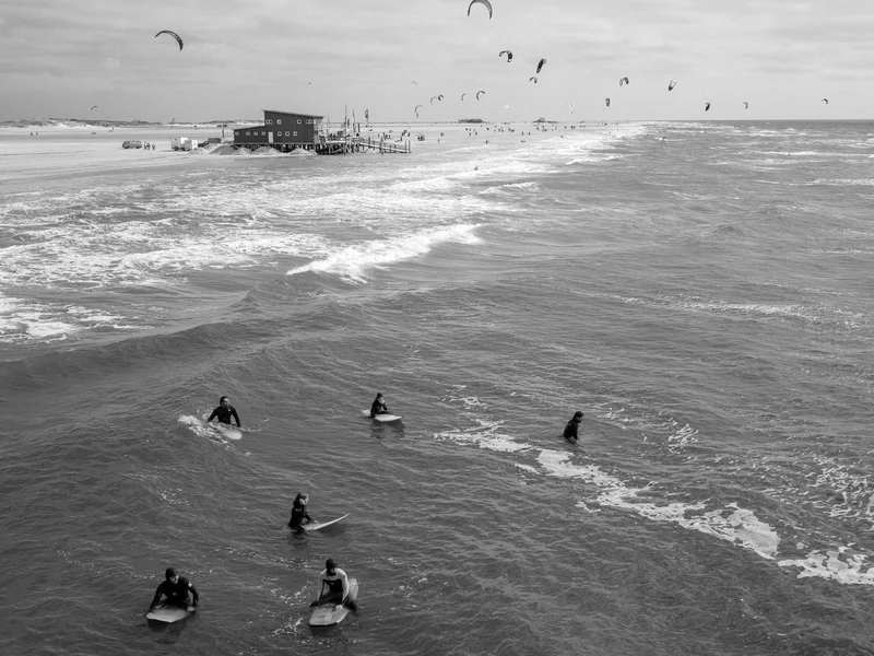 Surfers on the water with a beach in the distance and kites in the sky.