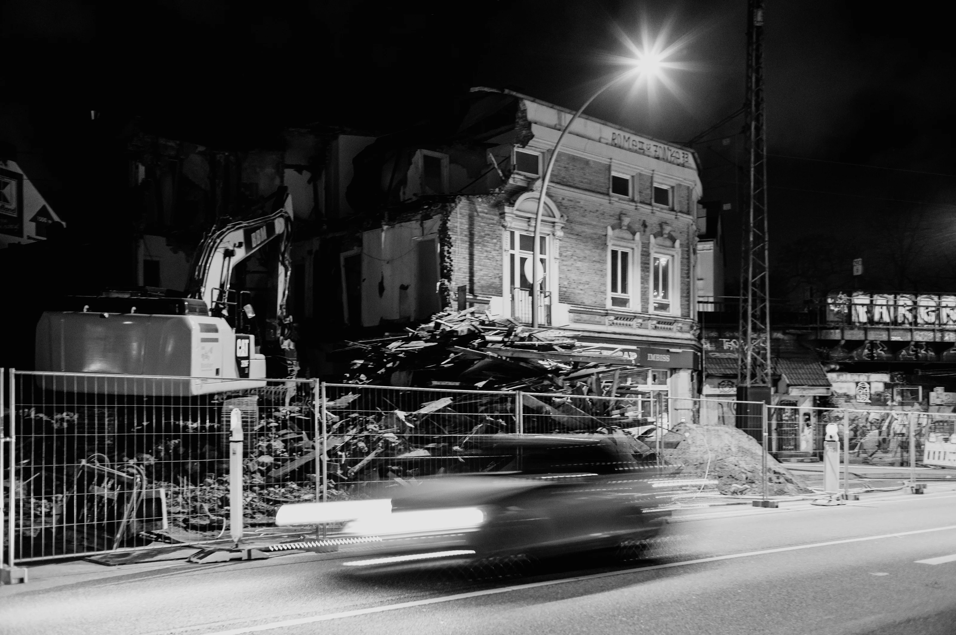 Demolition site with excavator and rubble beside a blurred moving car at night.