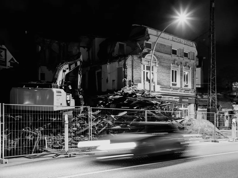Demolition site with excavator and rubble beside a blurred moving car at night.