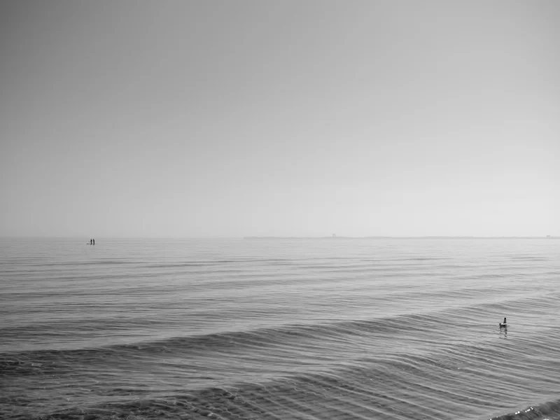 A minimalist black and white photo of a calm sea with a distant paddleboarder on the horizon.