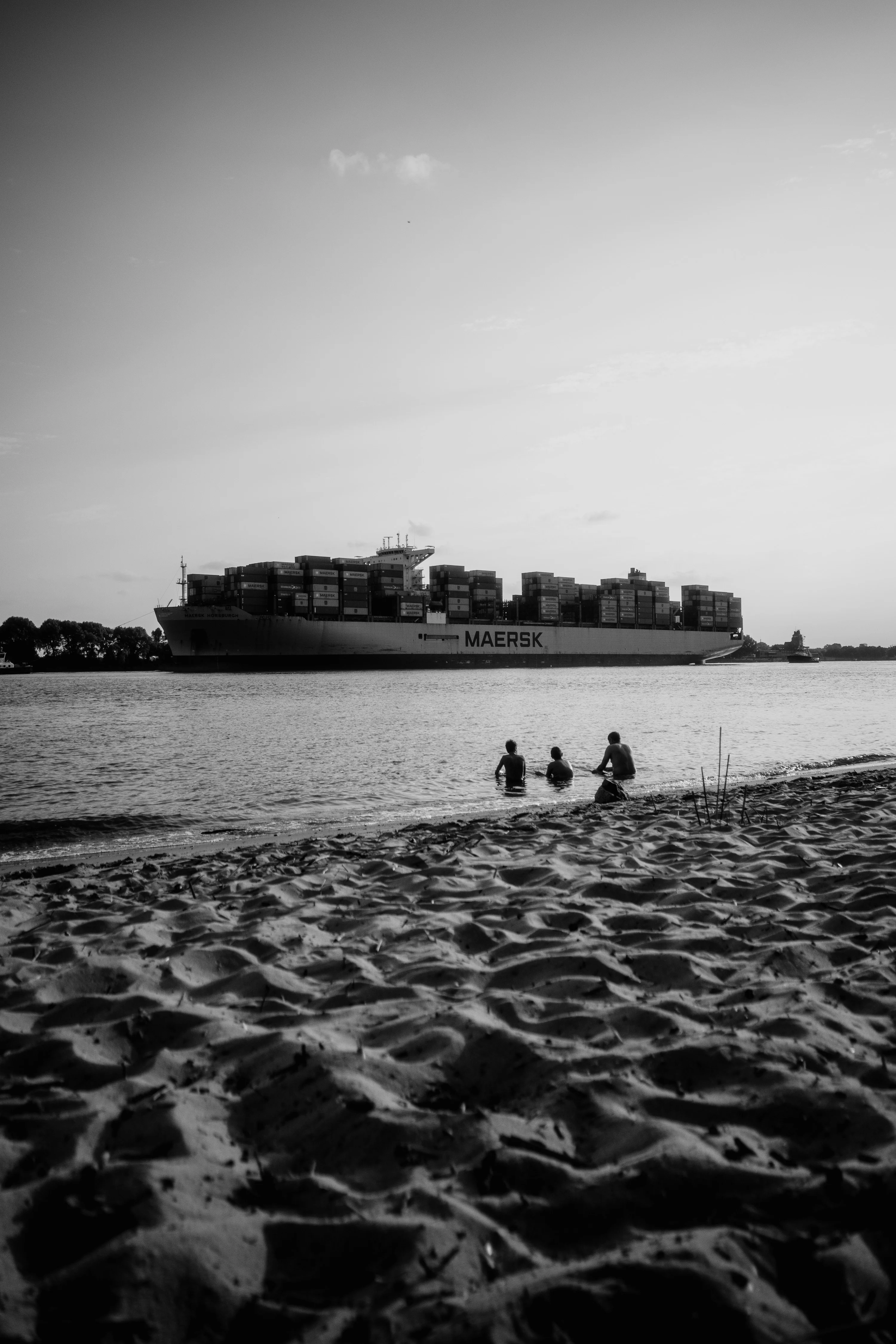 Three people sitting on a sandy beach with a large container ship passing by on the water.