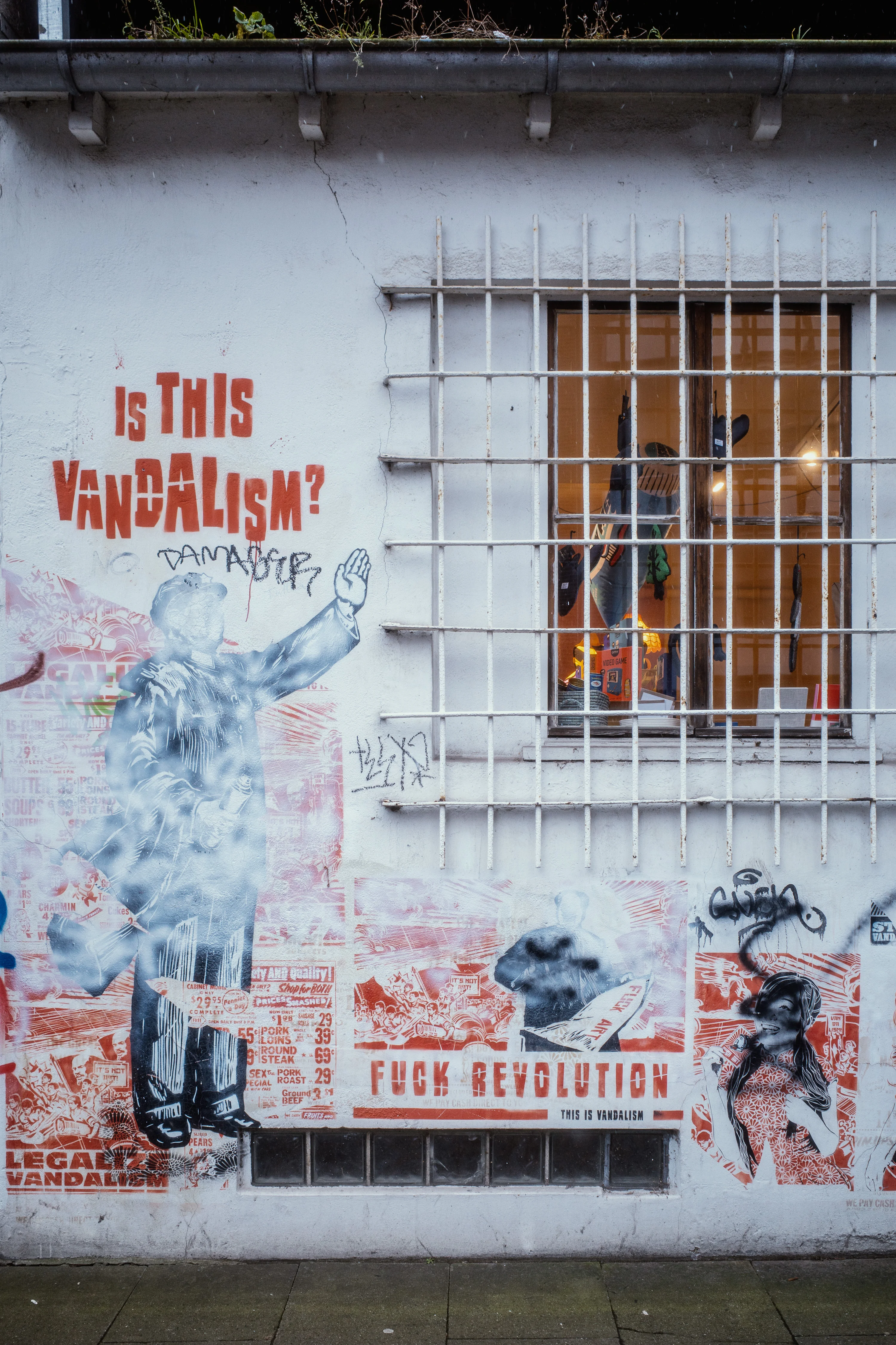 Street art featuring a question about vandalism alongside various posters on a wall with a barred window.