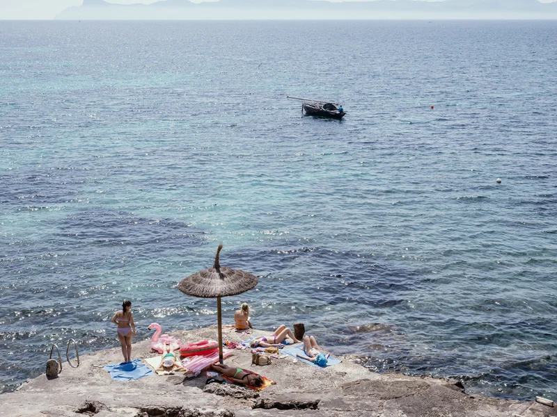 People relaxing with inflatables on a rocky beach by the sea under a straw parasol.
