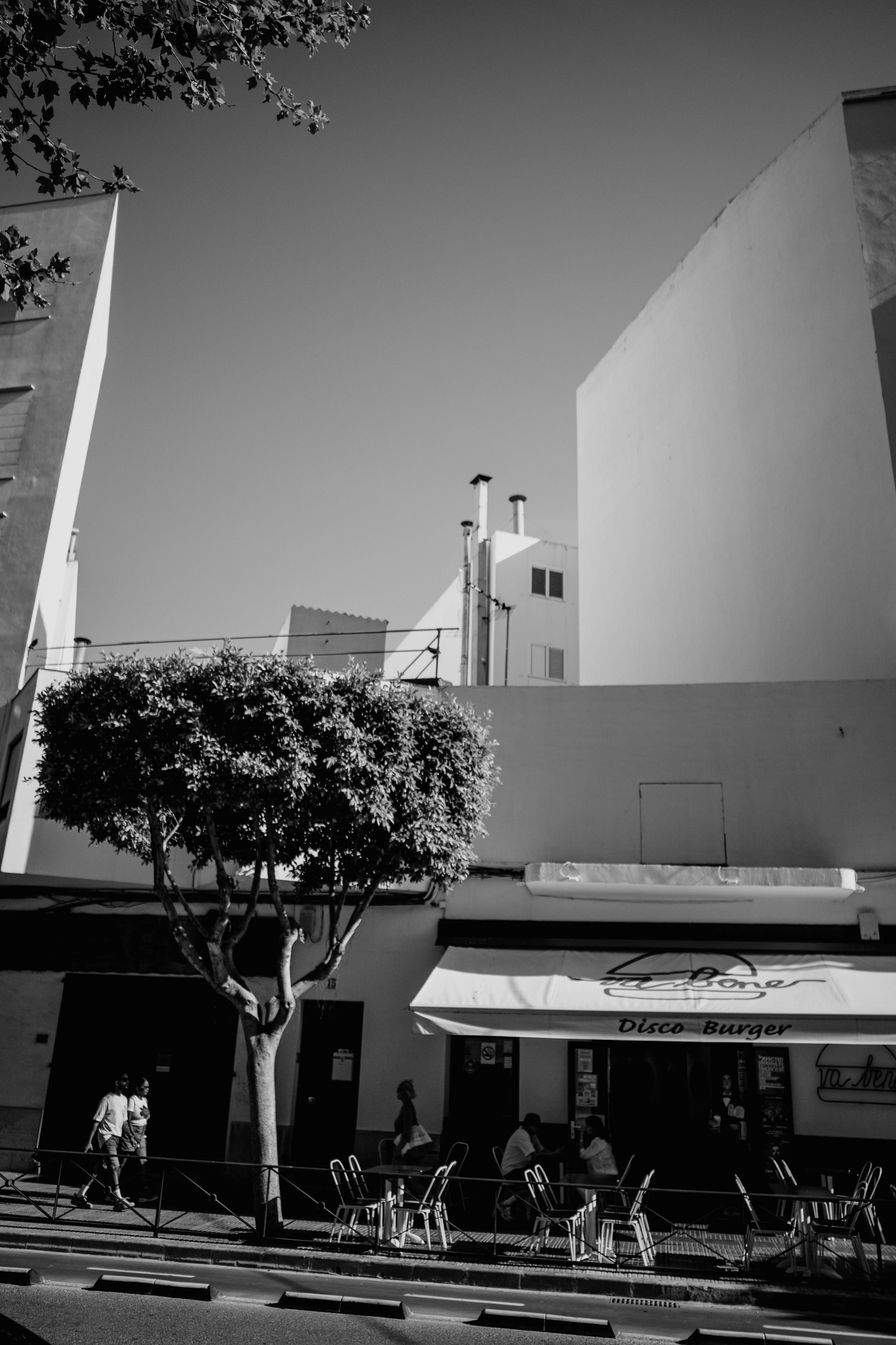 A black and white street scene with a tree in front of a building labeled 'Disco Burger' and people walking and sitting at tables.