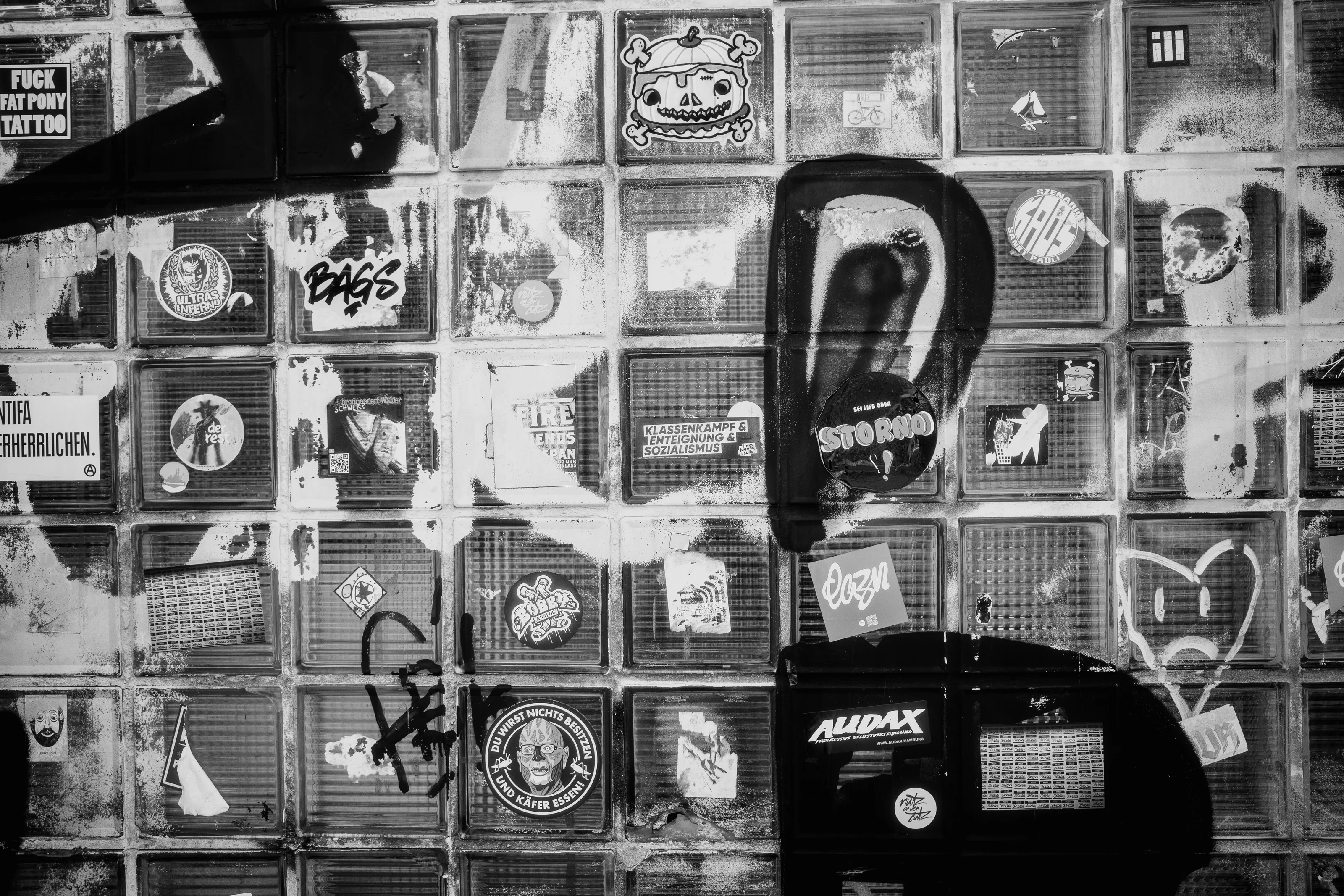 A monochrome wall with various stickers and graffiti art on glass blocks.