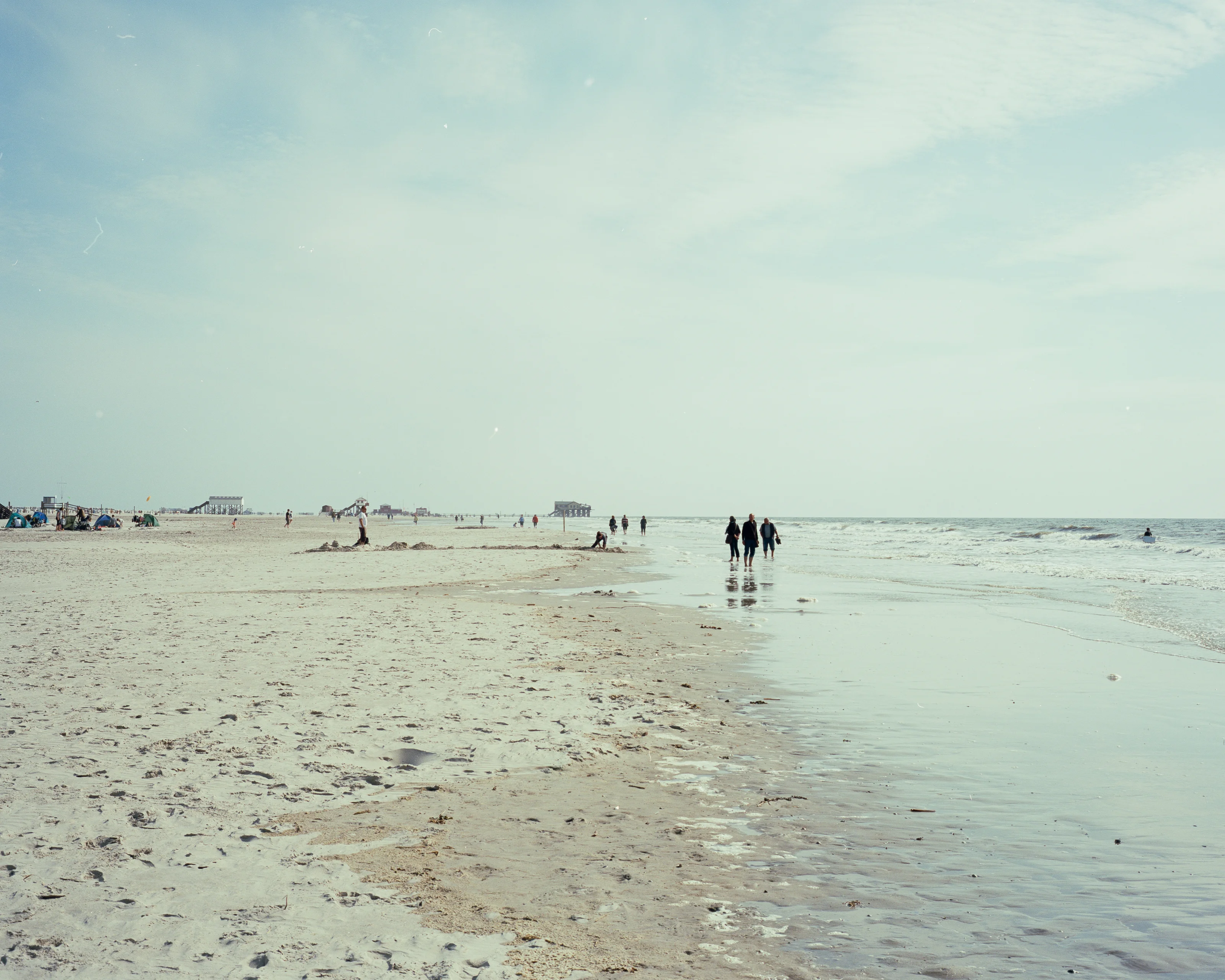 A wide sandy beach with people walking along the shoreline under a cloudy sky.