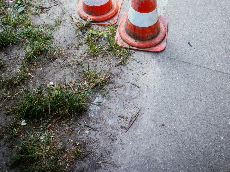 Two orange and white traffic cones on a patch of dirt and grass near a paved path and bicycle.