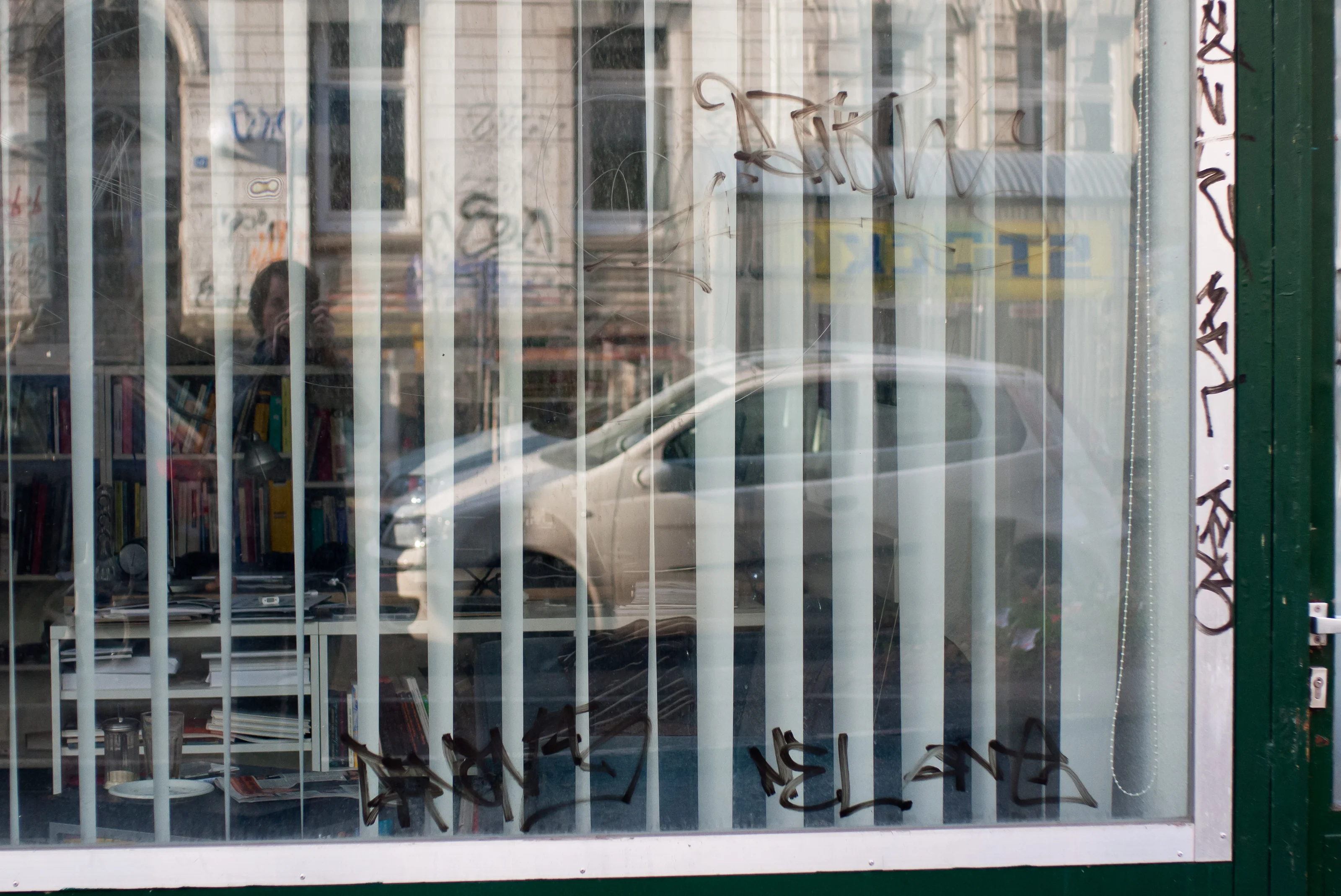 A car reflected in a glass window with vertical blinds and graffiti.