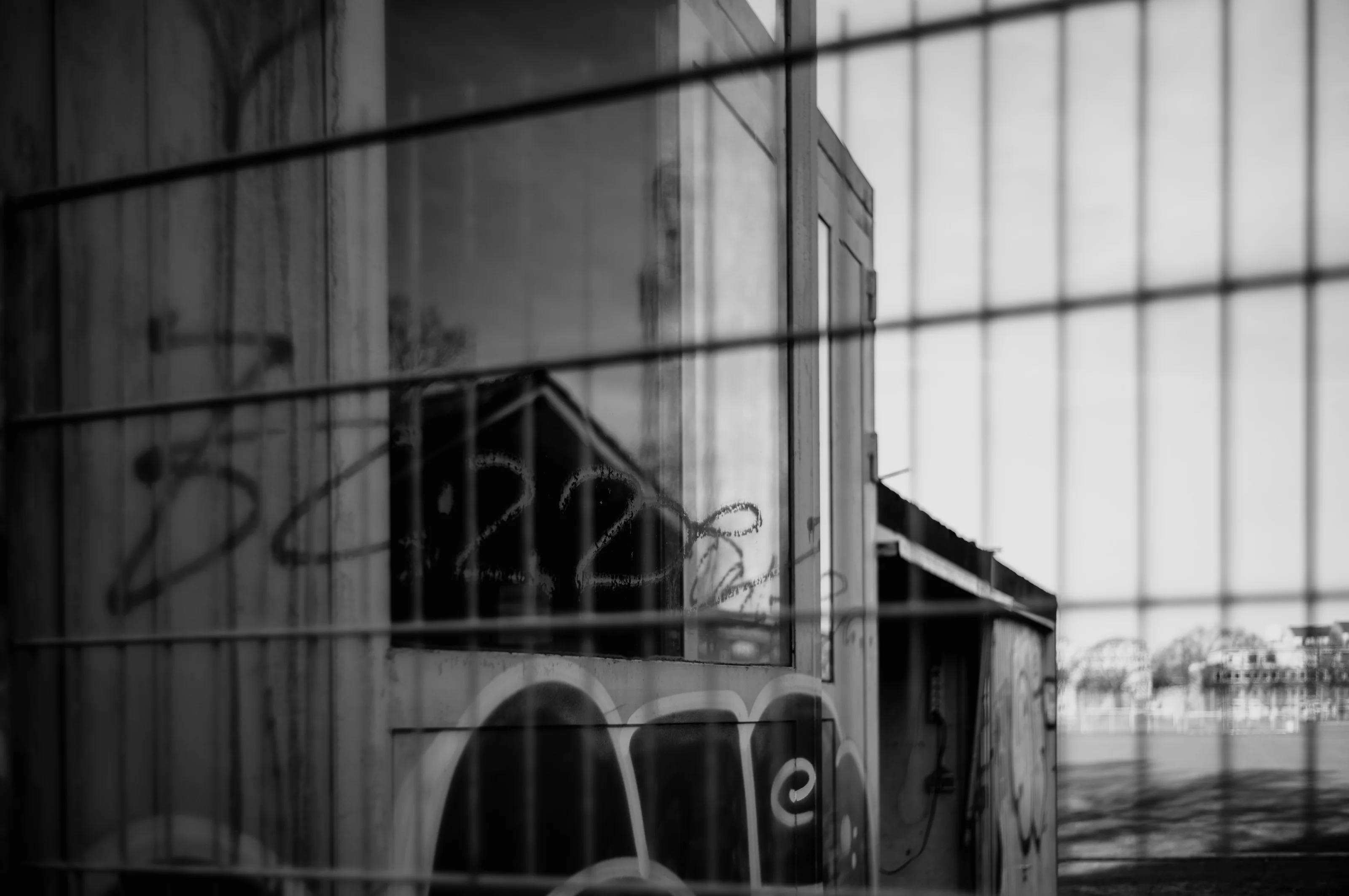 A monochrome photograph of a building with graffiti, viewed through a wire fence.