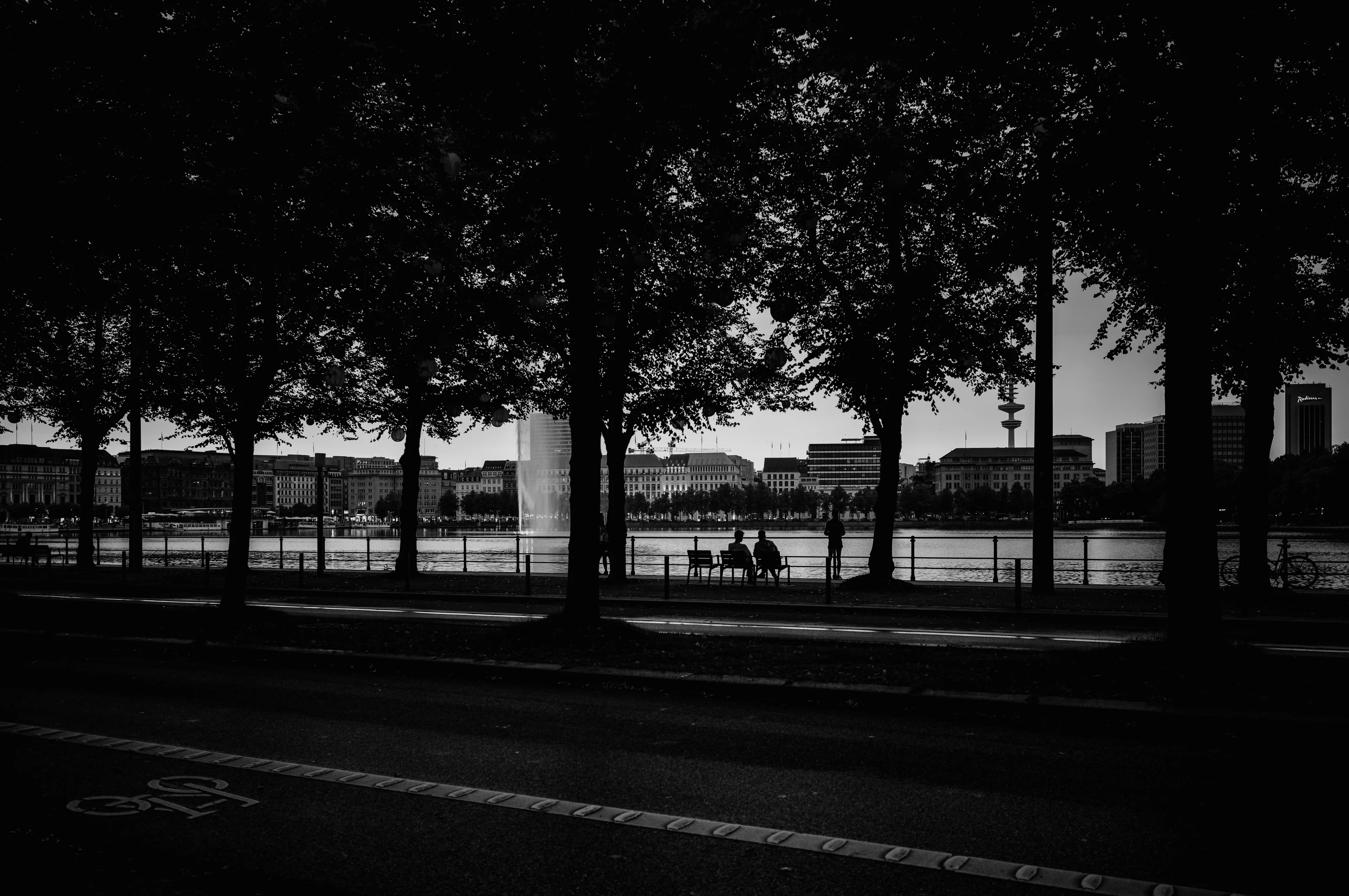 Silhouettes of people sitting and standing by a riverside, framed by trees with buildings in the background.