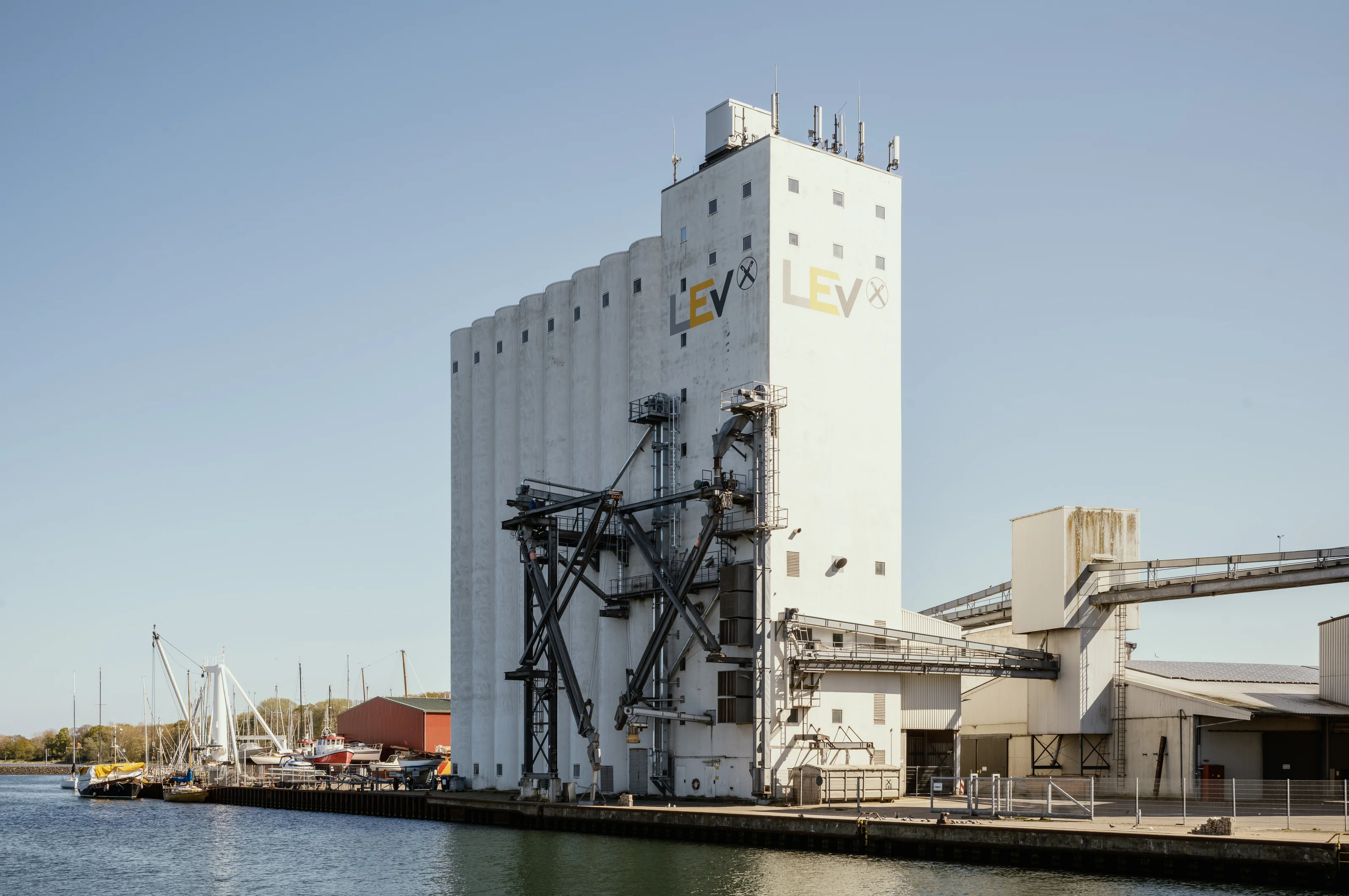 Large industrial building with pipes by a waterfront under a clear blue sky.