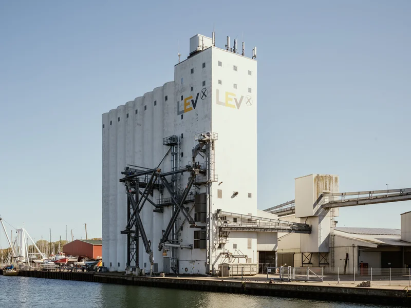 Large industrial building with pipes by a waterfront under a clear blue sky.