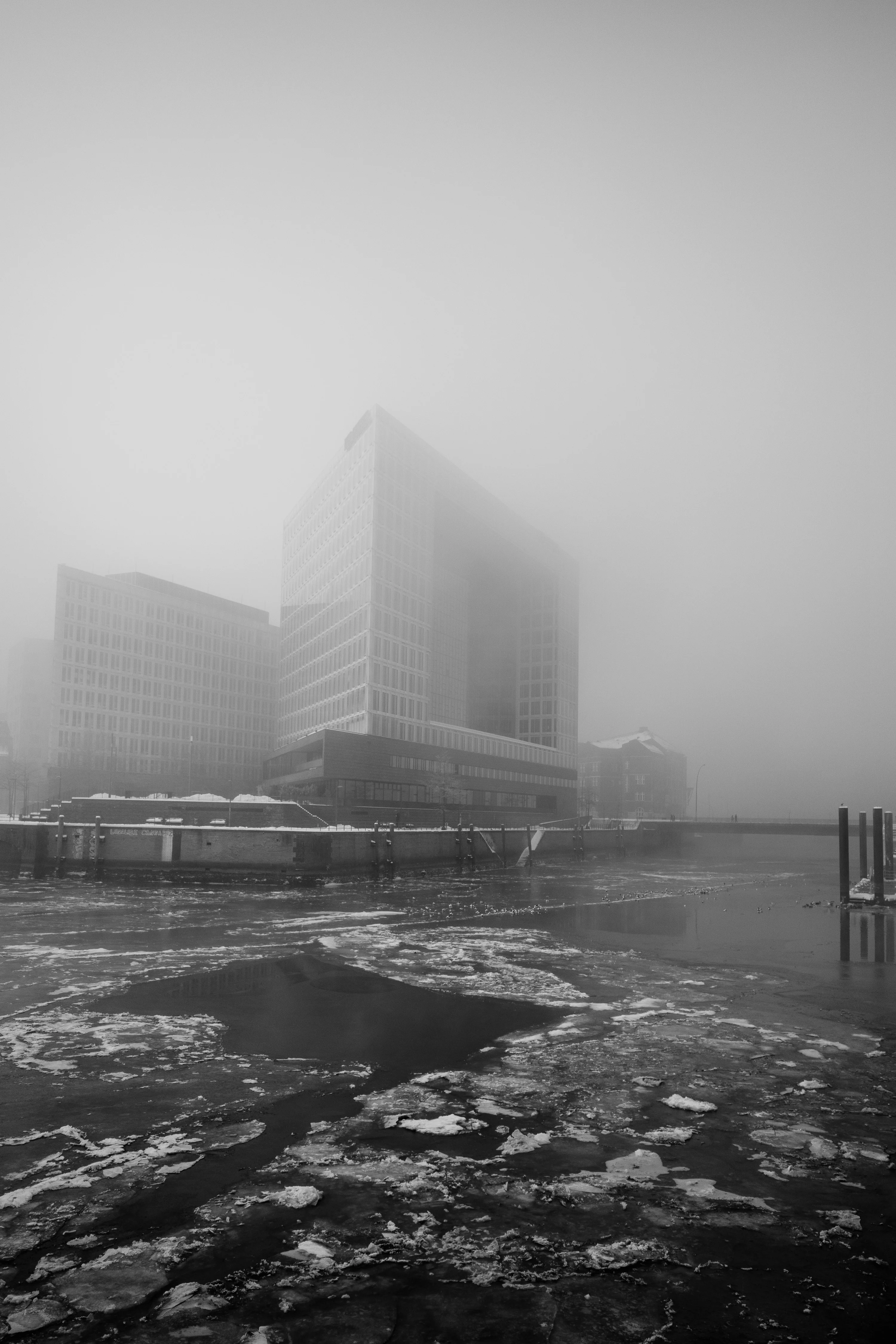 A tall modern building partially obscured by fog looms over a frozen body of water.